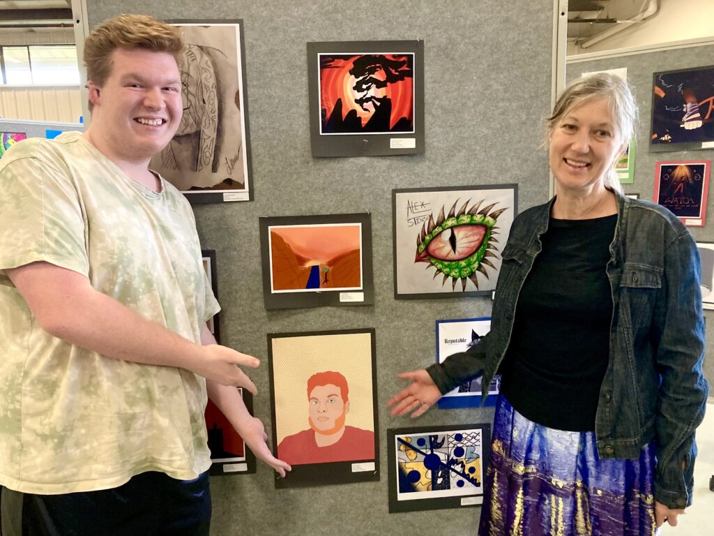 A smiling young man and woman stand in front of an art display, pointing to a framed self-portrait on the wall.