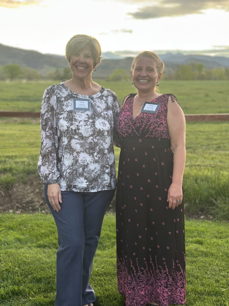 Two women stand together outdoors, smiling, wearing name tags, with a scenic green field and mountains in the background.