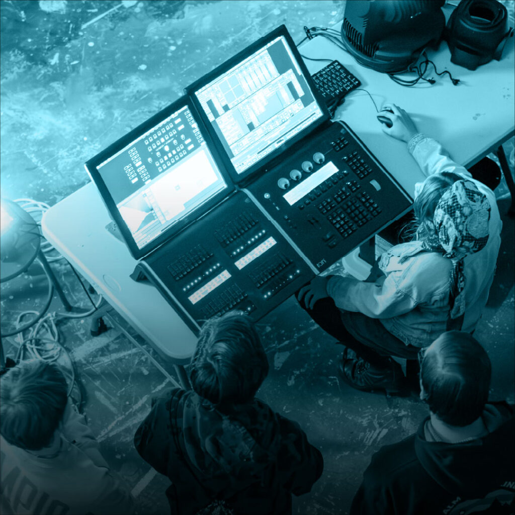 A person operates a lighting control desk with two monitors, while others observe from behind.