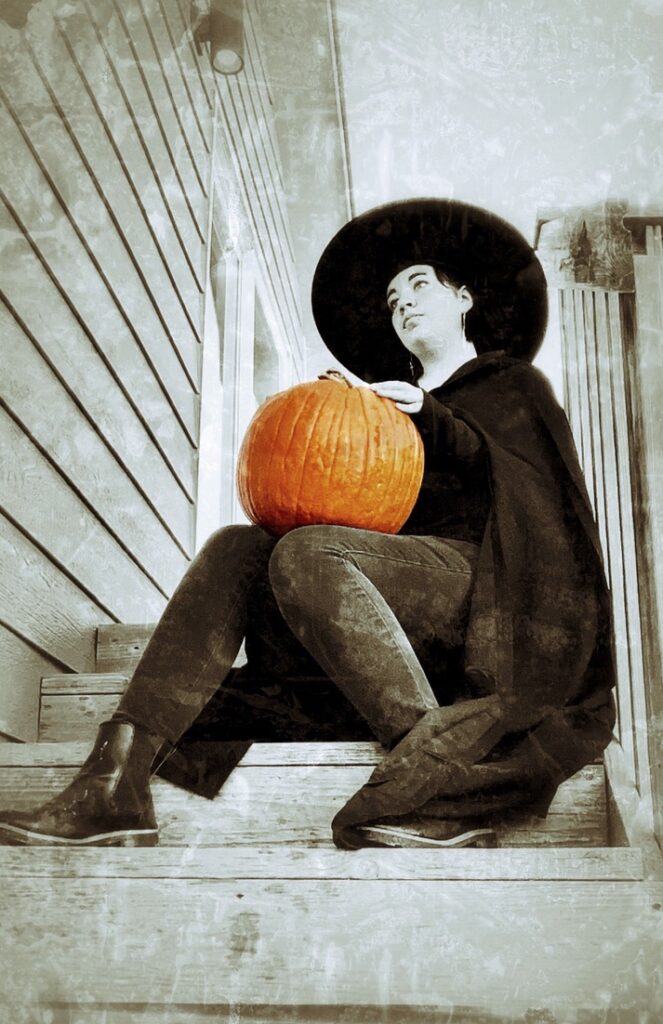 A person dressed as a witch sits on wooden steps, holding a pumpkin and looking thoughtfully upward.