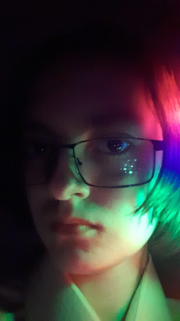A close-up portrait of a person with glasses, illuminated by colorful lights in a dark background.