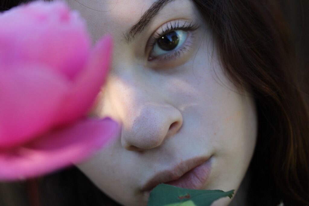 A close-up of a person's face partially obscured by a pink flower, with soft natural lighting highlighting features.
