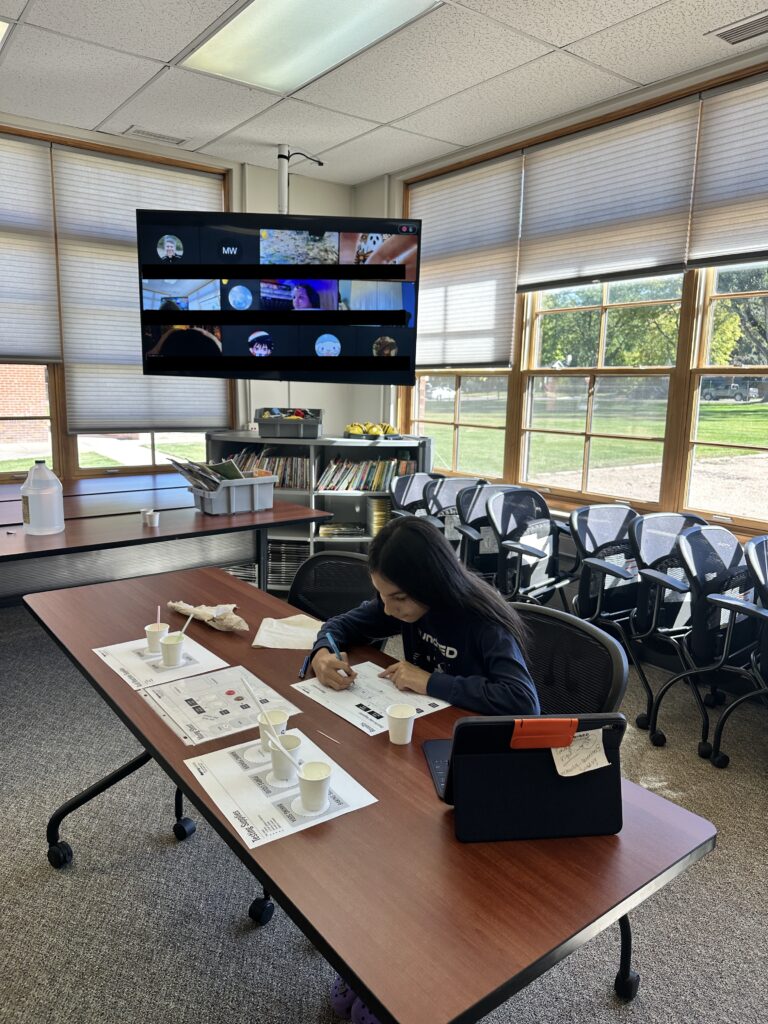 A student works at a table with papers and cups, while a TV displays a virtual meeting in a classroom setting.