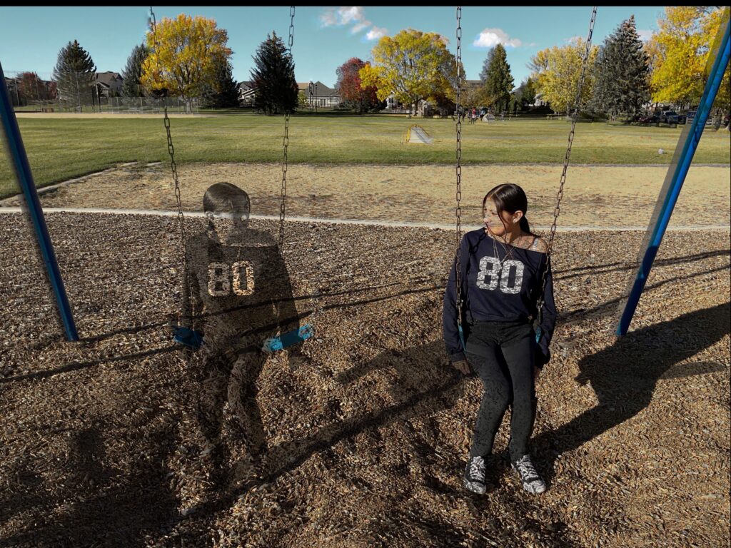 A girl sits on a swing in a park, with a ghostly image of another child also on the swing beside her.