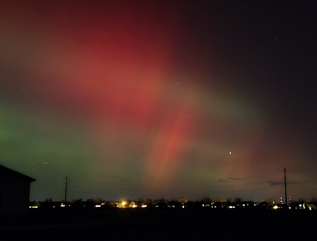 Vibrant auroras in red and green hues illuminate the night sky above a dark landscape with distant lights.