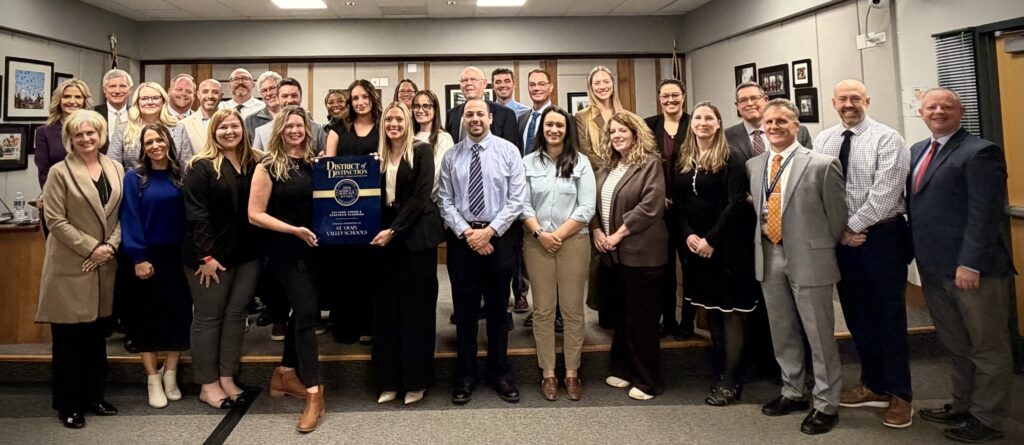 A group photo of staff members holding a "District of Distinction" award in a conference room setting.