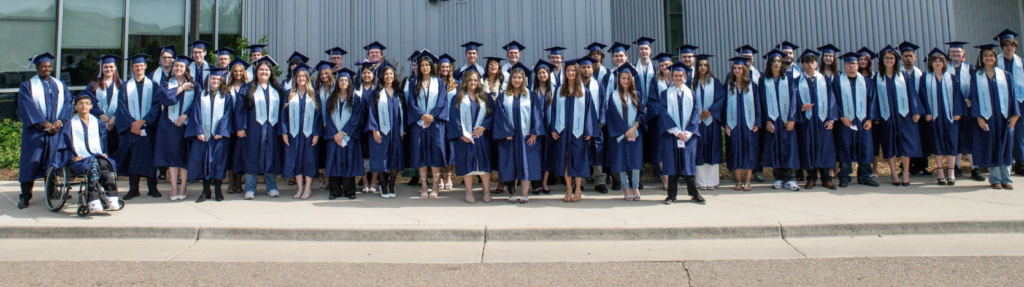 A large group of graduates in blue gowns and caps poses together outside, smiling and celebrating their achievement.