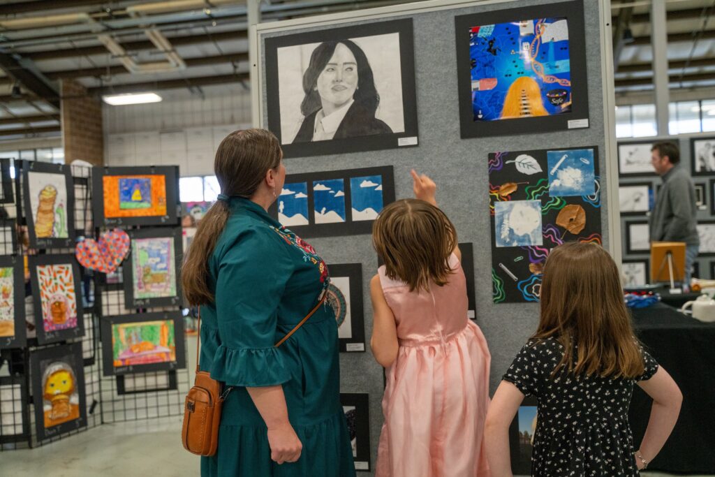A woman and two girls admire artwork at an exhibition, pointing at a portrait and discussing the displayed pieces.