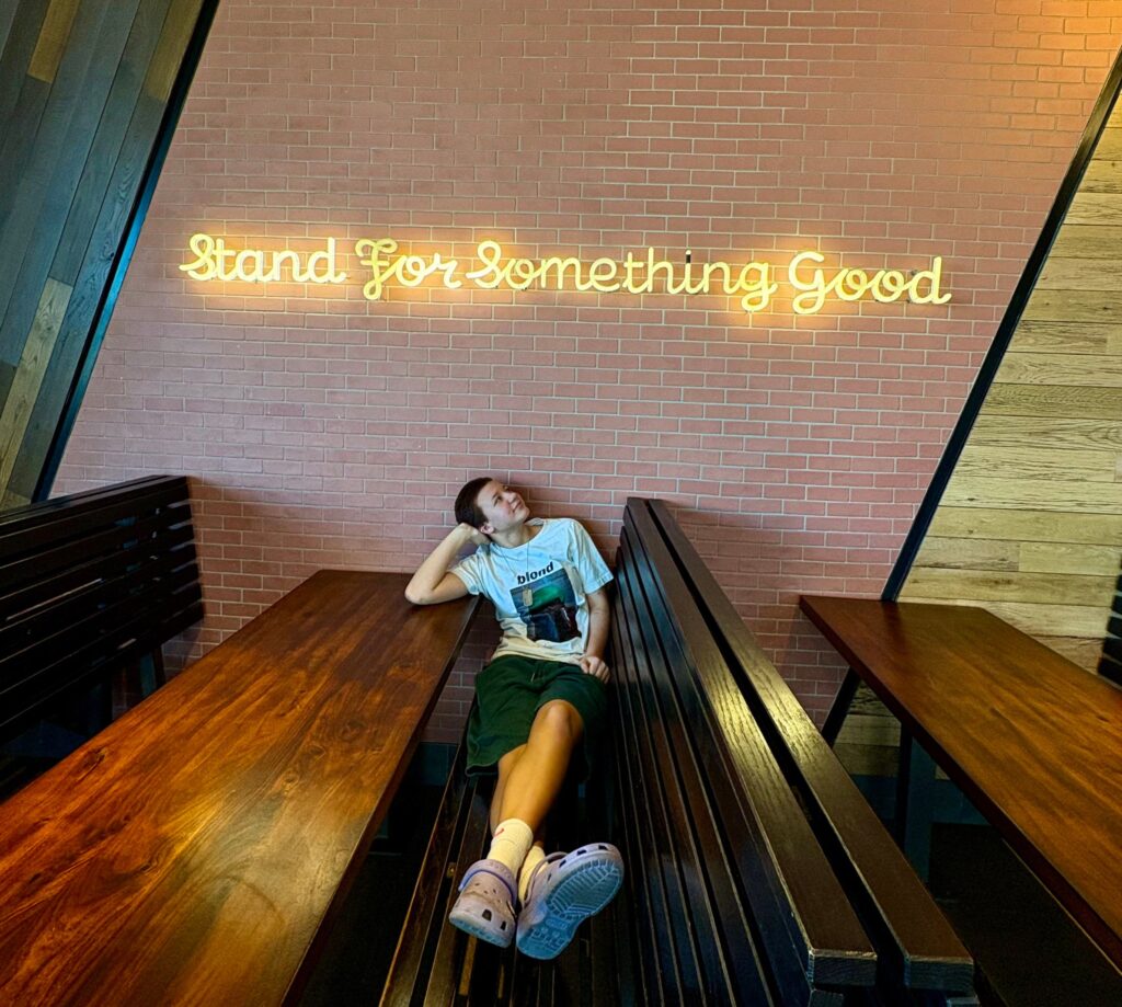 A person relaxes on a wooden bench under a neon sign that says "Stand For Something Good" on a brick wall.